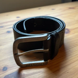 Svart skärp från Jack & Jones i skinn - Snyggt brunt bälte från Jack & Jones i äkta skinn med präglad logga längs remmen. Kraftigt metallspänne med märkets namn ingraverat. Perfekt accessoar för att lyfta din outfit med en stilren och klassisk touch. Bältet är dock ganska slitet, men i övrigt väldigt bra skick