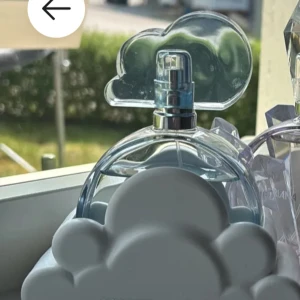 Ariana Grande Cloud parfym - Superfin parfym från Ariana Grande i den ikoniska Cloud-flaskan. Flaskan är genomskinlig med blå toner och har ett lock i form av ett moln. Parfymen står i en söt vit molnformad hållare som gör den extra dekorativ på hyllan.