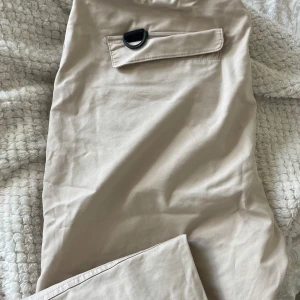 Beiga cargopants från Bershka  - Säljer ett par beiga cargopants från L1K3 med flera stora fickor, svarta detaljer och snörning i midjan. Byxorna har elastisk midja och dragkedjor på benen. 