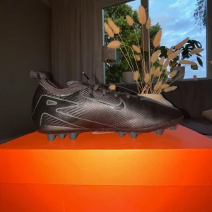 Svarta Nike fotbollsskor med dobbar - Snygga svarta Nike fotbollsskor ZM vapor 16-pro med diskreta silverdetaljer och klassisk swoosh på sidan. Skorna har snörning och en lätt, syntetisk ovandel med mönstrad yta för bättre bollkontroll. Utrustade med fasta dobbar för spel på gräs. Perfekta för dig som vill sticka ut på planen.