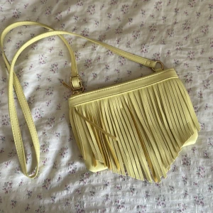 Gul axelväska med fransar - Såååå snygg ljusgul väska med långa fransar framtill och justerbar axelrem. Väskan har en mjuk, slät yta och stängs med dragkedja!