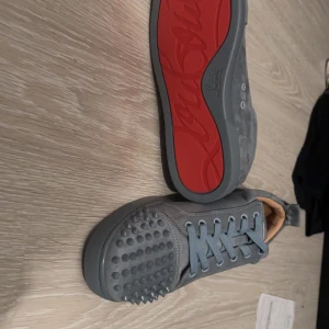 Grå Christian Louboutin sneakers med nitar - Snygga grå sneakers från Christian Louboutin i mocka med ikoniska röda sulor och coola nitar över tån. Skorna har snörning och rund tå, samt en lyxig känsla med läderfoder. Perfekta för dig som vill sticka ut med en edgy och exklusiv stil.