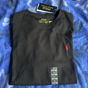 Svart t-shirt från Polo Ralph Lauren - Svart t-shirt från Polo Ralph Lauren med klassisk rund hals och ikonisk röd broderad logga på bröstet. Tillverkad i mjuk bomull som känns skön mot huden. Enkel och stilren design som passar perfekt till jeans eller shorts.