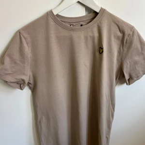 Beige t-shirt från Lyle & Scott - Snygg beige t-shirt från Lyle & Scott med klassisk gul logga på bröstet. T-shirten har rund halsringning och korta ärmar. 