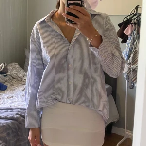 Randig blåvit skjorta i bomull - Klassisk skjorta med blå och vita ränder, oversized passform och långa ärmar. Skjortan har knappar framtill och bred krage, perfekt att styla både instoppad och lös. Tillverkad i mjuk bomull som känns skön mot huden. En liten sminkfläck fram där av priset.