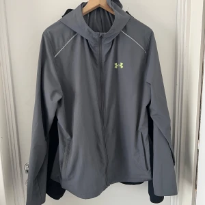 Grå vindjacka från Under Armour - Snygg grå vindjacka från Under Armour med huva och dragkedja framtill. Jackan har reflekterande detaljer och logga i neongult både fram och bak. Perfekt för blåsiga dagar och sportiga outfits. 