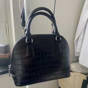 Svart handväska - Snygg svart handväska i skinnimitation. Väskan har rundad form, dubbla handtag, axelrem och dragkedja upptill. Insidan har ett fack med dragkedja för småsaker. Det långa axelbandet har gått sönder på ett litet ställe!