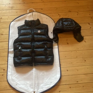 Svart dunväst från Moncler med huva - Svart, glansig dunväst från Moncler med avtagbar huva och två dragkedjefickor. Klassisk Moncler-logga på bröstet och coolt serietryck på insidan. Perfekt för lager-på-lager och streetstyle.
