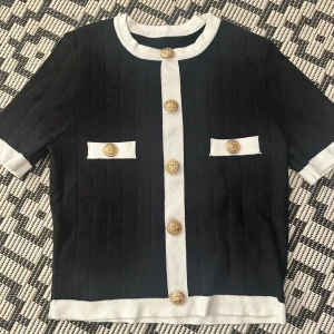 Svartvit kofta med guldknappar från Cider - Stilren svart kofta med vita detaljer från Cider. Kortärmad modell med ribbad struktur, dekorativa guldknappar framtill och vid fickorna. Materialmix av viskos, polyester och polyamid. Perfekt för dig som gillar en klassisk men trendig look.