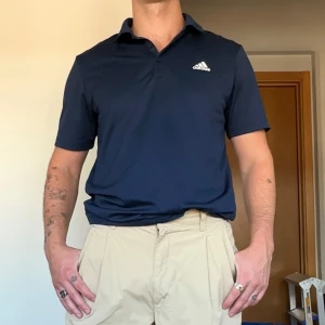 Mörkblå pikétröja från Adidas - Snygg mörkblå pikétröja från Adidas med klassisk krage och korta ärmar i nyskick. Tröjan har Adidas-logga på bröstet och är tillverkad i ett mjukt och lätt material som passar perfekt till sommaren. Enkel och stilren design som funkar till många outfits.