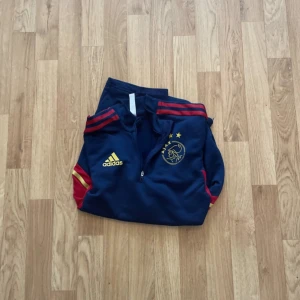 Ajax x Adidas blå långärmad träningströja - Snygg långärmad träningströja från Adidas och Ajax i marinblått med röda och gula detaljer. Tröjan har half zip, gult Ajax- och Adidas-tryck på bröstet samt röda ränder på axlarna. Tillverkad i polyester, perfekt för fotbollsträning.