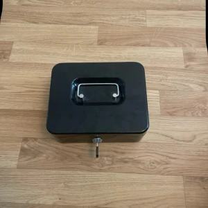 Svart plånboksbox med handtag - En svart plånboksbox i metall med praktiskt handtag på locket och lås framtill. Insidan har ett löstagbart fack för mynt och sedlar. Perfekt för att hålla koll på pengar eller småprylar. Enkel och stilren design som funkar i många sammanhang.