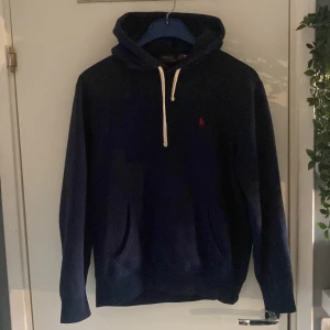 Mörkblå hoodie från Polo Ralph Lauren - Säljer en mörkblå hoodie från Polo Ralph Lauren med klassisk röd logga på bröstet. Tröjan har huva med vita snören och en stor ficka framtill. Perfekt för dig som gillar stilren och enkel design.