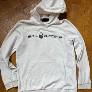 Vit hoodie från Sail Racing - Vit hoodie från Sail Racing med svart logga tryckt på bröstet. Klassisk känguruficka framtill, ribbade muddar och huva. Mjuk insida i bomullsblandning, perfekt för chill dagar eller när du vill ha en clean look.