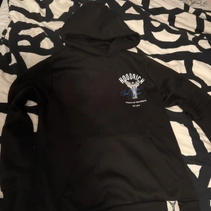 Svart HOODRICH hoodie med ängeltryck - Svart hoodie från HOODRICH med stor ängel och texten 'From Nothing To Something' i blått och vitt på ryggen. Liten logga och motiv på bröstet. Klassisk känguruficka och huva. Perfekt streetwear-vibe och riktigt snygg detalj på ryggen.liten skada på armen men syns inte så mycket 