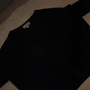 Svart kofta från H&M, storlek L - En klassisk svart kofta från H&M i storlek L. Koftan har knappar framtill och lång ärm, perfekt att slänga över en t-shirt eller linne. Enkel och stilren design som funkar till det mesta.