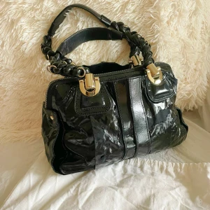 Bay patent Leather handväska från Chloé - Säljer en svart handväska från Chloé i glansigt lackat skinn med flätat handtag och snygga guldfärgade metalldetaljer. Väskan har dragkedja upptill och innerfack med Chloé-logga. Perfekt för dig som vill ha en statementväska med lyxig känsla. 23cm x34cm x10cm. 