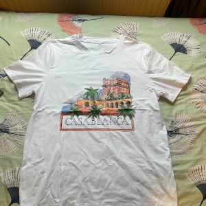 Vit Casablanca t-shirt med tryck - Vit t-shirt från Casablanca med ett stort färgglatt tryck på framsidan som visar en byggnad, palmer och texten 'CASABLANCA'. Klassisk rund hals och korta ärmar. Perfekt för dig som gillar snygga prints och streetstyle.