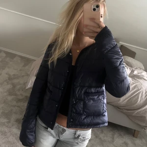 Mörkblå pufferjacka med fickor - Säljer en mörkblå pufferjacka med stora fickor framtill och tryckknappar. Jackan är kortare i modellen och har en clean look utan synliga loggor. Perfekt för dig som vill ha en enkel men snygg jacka till höst. 