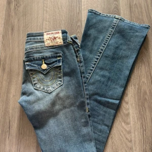 True Religion Joey flare jeans blå - Säljer ett par blå True Religion Joey jeans med låg midja och flare-ben. Klassiska kontrastsömmar, snygga detaljer på bakfickorna och guldiga knappar. Jeansen är i denim med dragkedja och har en riktigt ikonisk 2000-tals vibe.