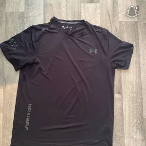 Svart Under Armour t-shirt i polyester - Snygg svart t-shirt från Under Armour med regnbågsfärgad logga på bröstet och textdetaljer på ärmen och sidan. Tillverkad i mjuk och lätt polyester, perfekt för träning eller chill. Klassisk passform med korta ärmar och rund hals.