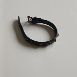 Svart armband med nitar Valentino  - Coolt svart armband med trekantiga silverfärgade nitar och spänne. Perfekt accessoar för att ge din outfit en edgy vibe. Justerbar storlek med flera hål för bästa passform. Ny pris 3500