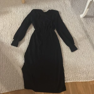 Svart långklänning med långa ärmar - En svart långklänning med långa ärmar och markerad midja. Klänningen har en elegant, figurnära passform och är tillverkad i ett tyg med subtil glans och struktur. Perfekt för dig som vill ha en stilren och klassisk look.