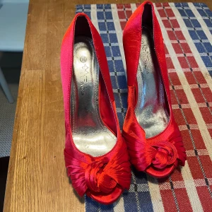 Röda pumps med rosett från HT Shoes - Snygga röda pumps i satin från HT Shoes, storlek 39. Skorna har öppen tå, hög klack och en dekorativ rosett framtill. Perfekta för dig som vill sticka ut med färg och elegans. Insidan är metallicfärgad och sulan har ett coolt mönster.