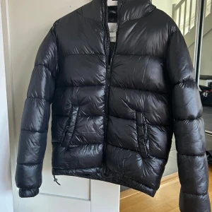 SVART ODIN JACKET - KILLE storlek 158-164 - Säljer en svart jacka från Grunt i storlek L (158-164). Köpt på Kids brand store. Jackan har glansig finish, huva, dragkedja och två snedställda fickor med knappdetalj. Perfekt för kalla dagar när du vill hålla stilen och värmen.