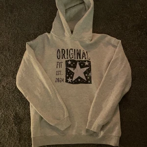 Scandi hoodie med stjärntryck - Snygg ljusgrå hoodie från Scandi med stor huva och tryck framtill. Trycket har en stor stjärna och texten 'Original Fit Est. 2024'. Mjuk och skön, perfekt för chill dagar. Klassisk passform och ribbade muddar vid ärmslut och midja.