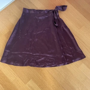Vinröd omlottkjol i satin - Snygg vinröd omlottkjol i glansigt satinmaterial med knyt i midjan. Kjolen har en lätt A-linjeform och är perfekt för dig som gillar en stilren och trendig look. Passar till både sneakers och klackar. Pris kan diskuteras💕