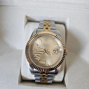 Tvåtonad klocka med gulddetaljer - Stilren klocka med tvåfärgad länk i silver och guld. Urtavlan är guldig med markerade index och datumfönster. Boetten har en räfflad guldfärgad kant som ger en lyxig känsla. Perfekt accessoar för dig som gillar klassisk och tidlös design.