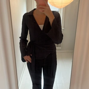 Svart omlottblus med knytband - Säljer en svart omlottblus med långa vida ärmar och snygg krage. Västen har ett brett knytband i midjan som har ett lätt, lite glansigt tyg. Väldrigt stilren. I storlek XS från Gina.