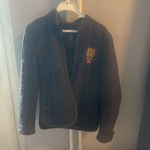 Svart quiltad jacka från Morris M - Snygg svart quiltad jacka från Morris i storlek medium. Jackan har broderad logga och brittisk flagga på bröstet, ståkrage och diskreta knappar vid ärmslut. Perfekt för dig som gillar klassisk och stilren look.