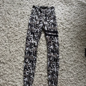 Stronger leggings med graffiti-mönster - Snygga leggings från Stronger med coolt graffiti-inspirerat mönster i svart, vitt och beige. Hög midja och tight passform, perfekta för träning eller chill. Materialet är stretchigt och mjukt för maximal komfort. Texten 'GIRL POWER' och 'SMILE' syns över hela byxan.