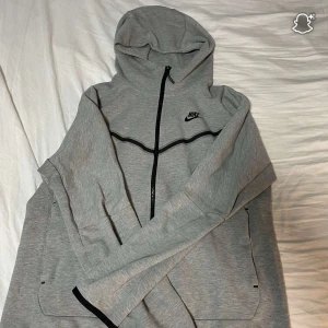 Grå Nike hoodie med dragkedja M - Snygg grå hoodie från Nike med svart dragkedja och svarta detaljer. Tröjan har huva, långa ärmar och två fickor framtill. Perfekt för chill dagar eller när du vill ha en sportig look. Klassisk Nike-logga på bröstet och stilren design.