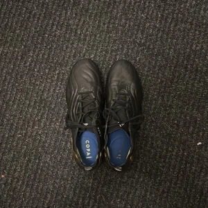 Adidas Copa Sense+ svarta fotbollsskor - Säljer ett par Adidas Copa Sense+ fotbollsskor i svart med snygga gulddetaljer på sidan och hälen. Skorna har en vågig design, snörning och är tillverkade i skinn för bästa känsla på planen. Perfekta för dig som vill sticka ut med stil på matchen.