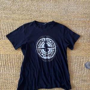 Svart Stone Island t-shirt med tryck - Svart t-shirt från Stone Island med stort vitt logotryck på bröstet. Klassisk rund hals och korta ärmar. Skön passform och mjukt material som känns nice mot huden. Perfekt för dig som gillar streetwear och vill sticka ut med en ikonisk look.