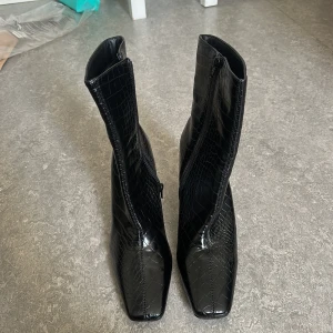 Nya Svarta boots - Nya Snygga svarta boots med krokopräglad yta och dragkedja på sidan. Skorna har spetsig tå, hög klack och är tillverkade i glansigt syntetmaterial. Aldrig använda, endast testade inomhus. 