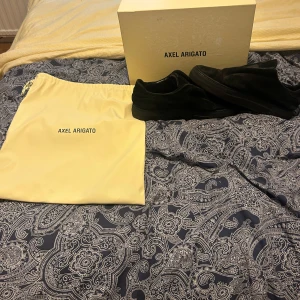 Svarta sneakers från Axel Arigato - Säljer ett par svarta sneakers från Axel Arigato i mocka med stilren design och platt sula. Skorna har en låg profil och kommer med originalkartong och dustbag. Perfekta för dig som gillar minimalistisk och clean stil.