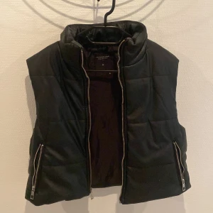 Svart dunväst från The Black Label - Snygg och modern svart quiltad dunväst från The Black Label Collection i modellen Blondie. Västen har hög krage, två fickor med dragkedja och stängs med en kraftig dragkedja framtill. Materialet är PU och polyester. Jättefint skick, inte använd.