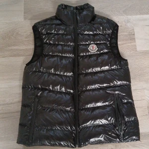 Svart dunväst från Moncler - Snygg svart dunväst från Moncler med glansig finish och klassisk logga på bröstet. Västen har hög krage, dragkedja framtill och två fickor med dragkedja. Perfekt att slänga över en hoodie eller tröja för extra värme.