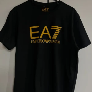 Svart EA7 Emporio Armani t-shirt - Svart t-shirt från EA7 Emporio Armani med stor gul logga framtill och gul detalj baktill. Klassisk rund hals och korta ärmar. Tillverkad i mjuk bomull för en skön känsla och snygg passform. Perfekt för dig som gillar stilrena och trendiga plagg.