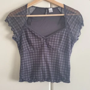 Rutig mesh topp från Divided - Supersöt rutig mesh topp från Divided i svart och grått. Toppen har korta, vågiga ärmar och en liten rosett vid bysten. Tunt, genomskinligt material och figurnära passform. Perfekt att styla med jeans eller kjol för en trendig look.