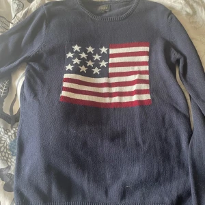 Mörkblå stickad tröja med USA-flagga - Mörkblå stickad tröja från Solid i storlek S. Tröjan har ett stort motiv av den amerikanska flaggan på bröstet och rund halsringning. Perfekt för dig som vill sticka ut med en cool och klassisk look.