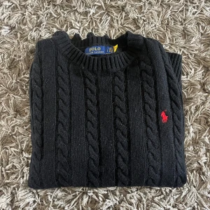 Svart kabelstickad tröja Polo Ralph Lauren - Kabelstickad svart tröja från Polo Ralph Lauren med klassisk rund halsringning och röd broderad logga på bröstet. Tröjan är i mjuk ull och har långa ärmar samt ribbade muddar för en snygg passform. Storlek L men känns som M