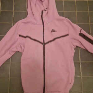 Rosa hoodie från Nike med dragkedja - Nike hoodie i ljusrosa med hel dragkedja och svart Nike-logga på bröstet. Snygg detalj med svart rand över bröstet och ärmficka med dragkedja. Huva med snörning och långa ärmar. Perfekt för chill eller träning.