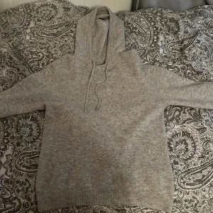 Grå hoodie med huva och snörning - Mysig grå hoodie med huva och snörning. Tröjan har lång ärm och ribbade muddar vid ärmslut och nederkant. Perfekt för chill dagar eller när du vill ha något bekvämt och stilrent. Enkel design som funkar till det mesta.