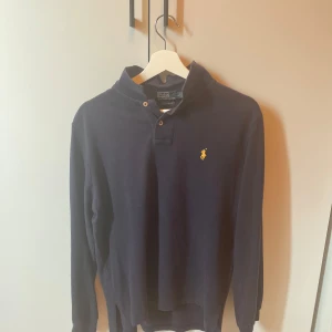 Mörkblå långärmad pikétröja Polo Ralph Lauren - Klassisk mörkblå långärmad pikétröja från Polo Ralph Lauren med gul broderad logga på bröstet. Tröjan har krage, två knappar vid halsen och är gjord i mjuk bomull. Perfekt för en stilren och avslappnad look. Skulle själv rekommendera den som en go present till en lillebrorsa som försöker fixa en go still 