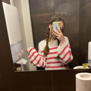 Randig stickad tröja från VILA - Superfin stickad tröja från VILA i vitt med rosa ränder. Tröjan har ribbad struktur, rund halsringning och långa ärmar. Tröjan är längre bak än fram och har lösa ärmar.